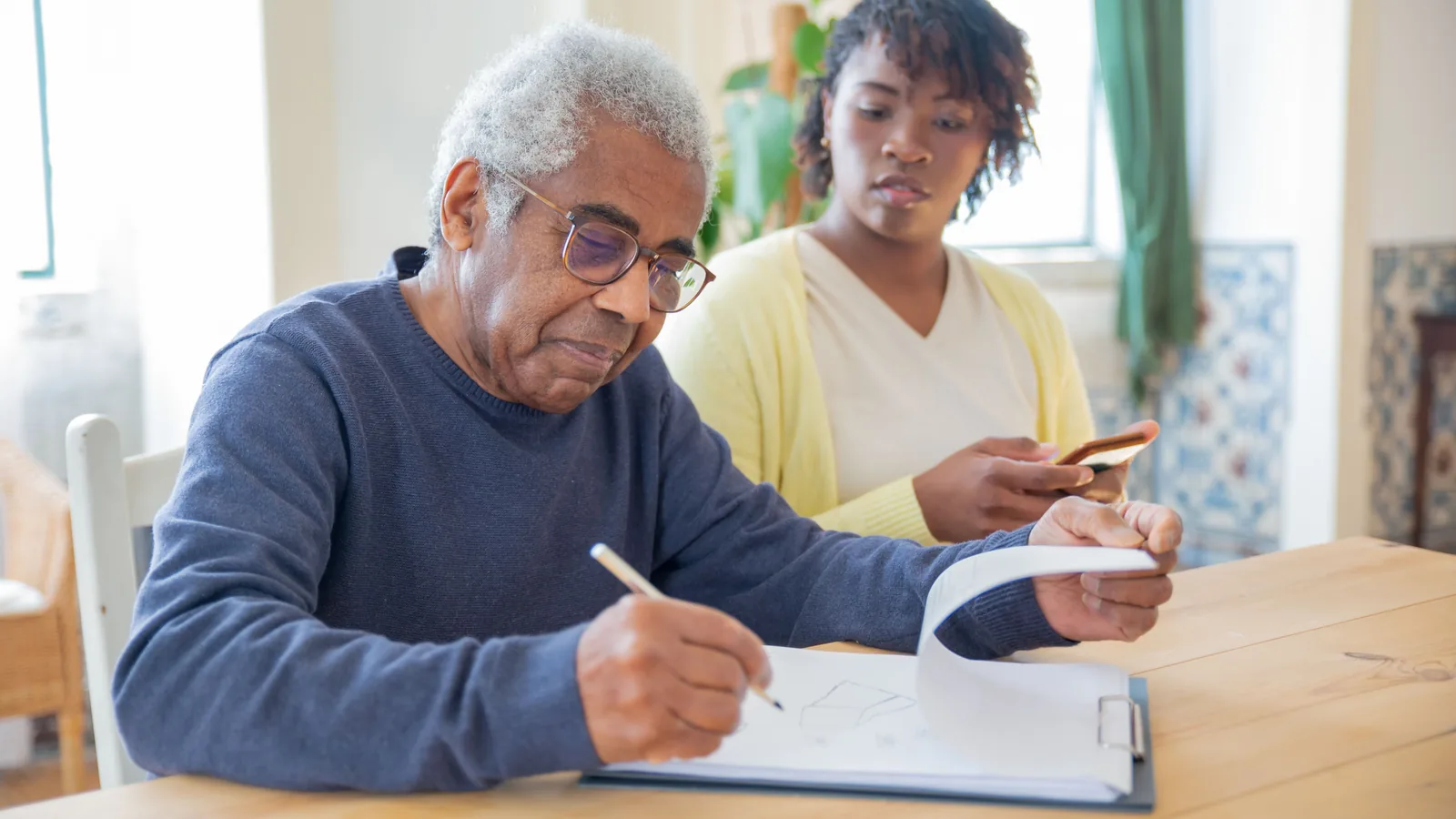Homme âgé rédigeant un document, accompagné d'une personne à ses côtés